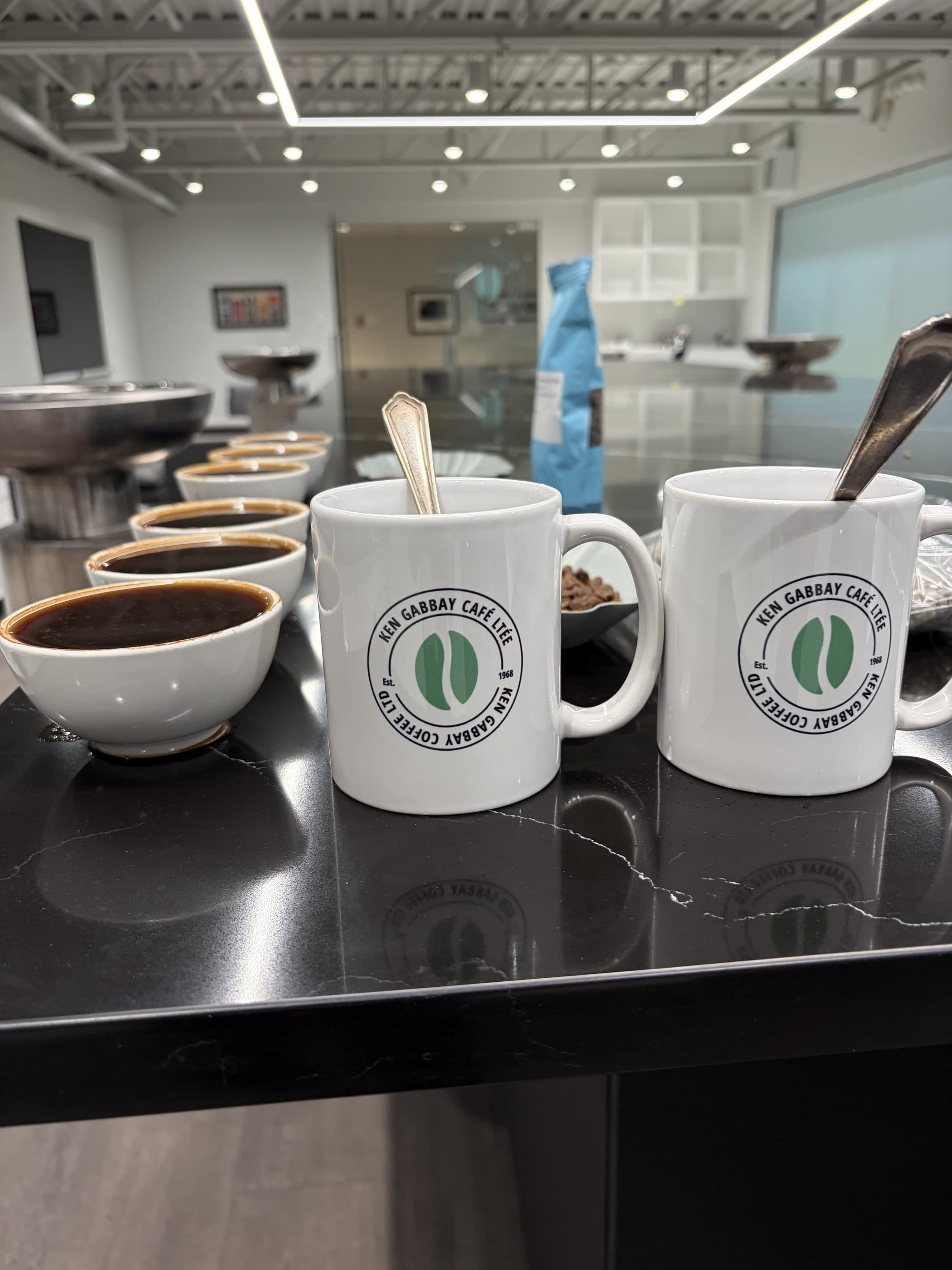 Une rangée de bols de dégustation blancs remplis de café infusé sur une table de pierre noire, avec au premier plan deux tasses imprimées du sceau Ken Gabbay Coffee, des cuillères posées dedans.